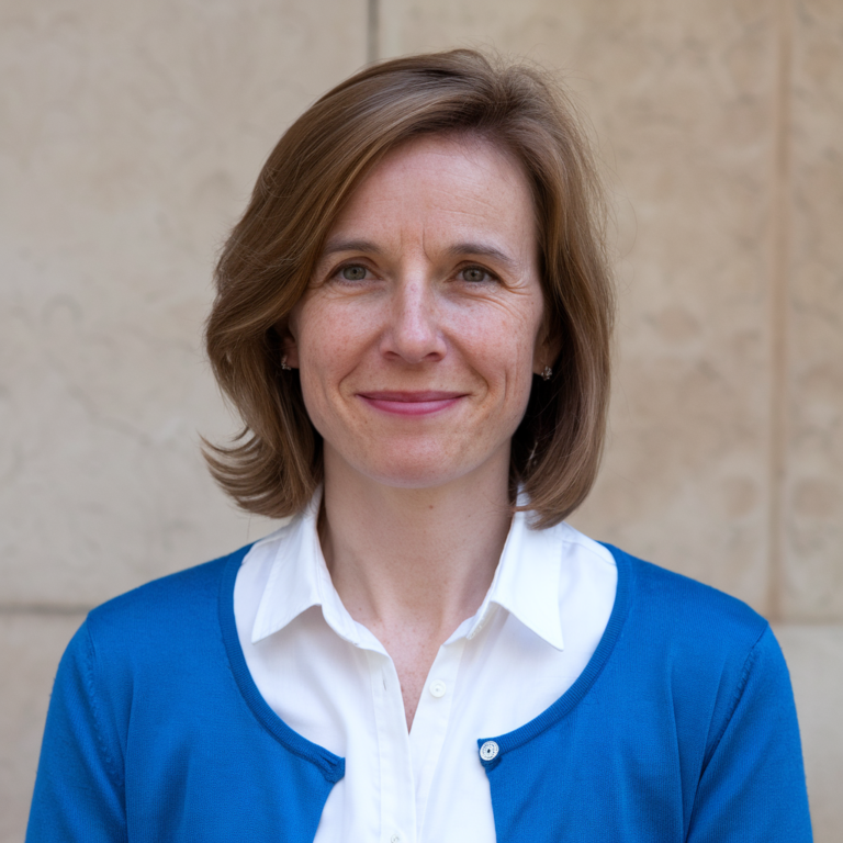 A woman with shoulder-length brown hair is smiling at the camera. She is wearing a white collared shirt and a blue cardigan. The background is a plain beige wall.